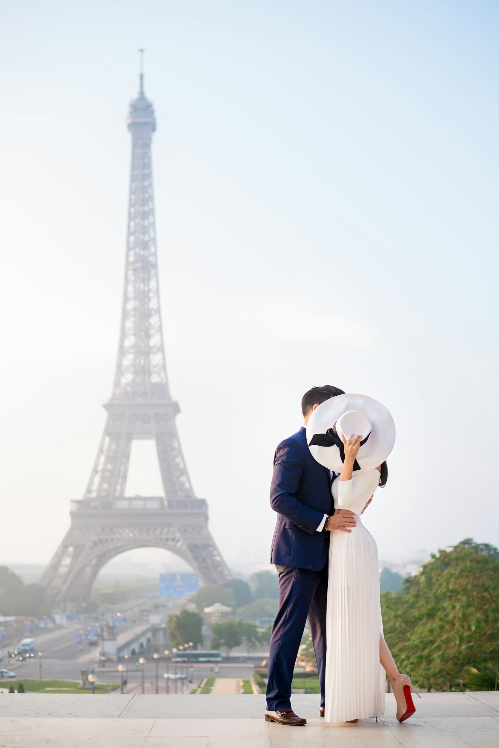A couple kissing behind a wide white hat on the Trocadéro, the Eiffel Tower in morning haze behind them