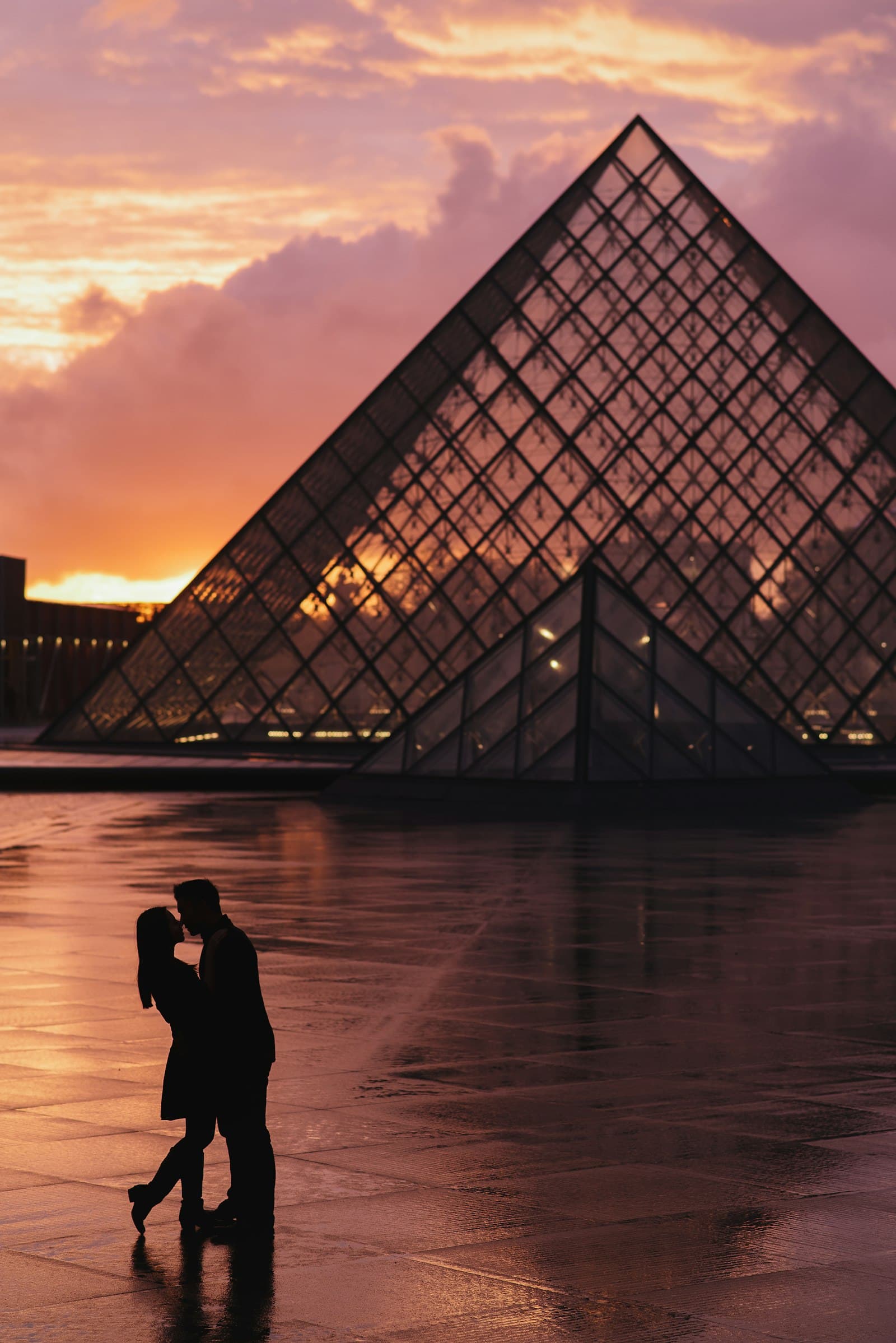 Silhouette of a couple kissing by the Louvre pyramid under a pink sky