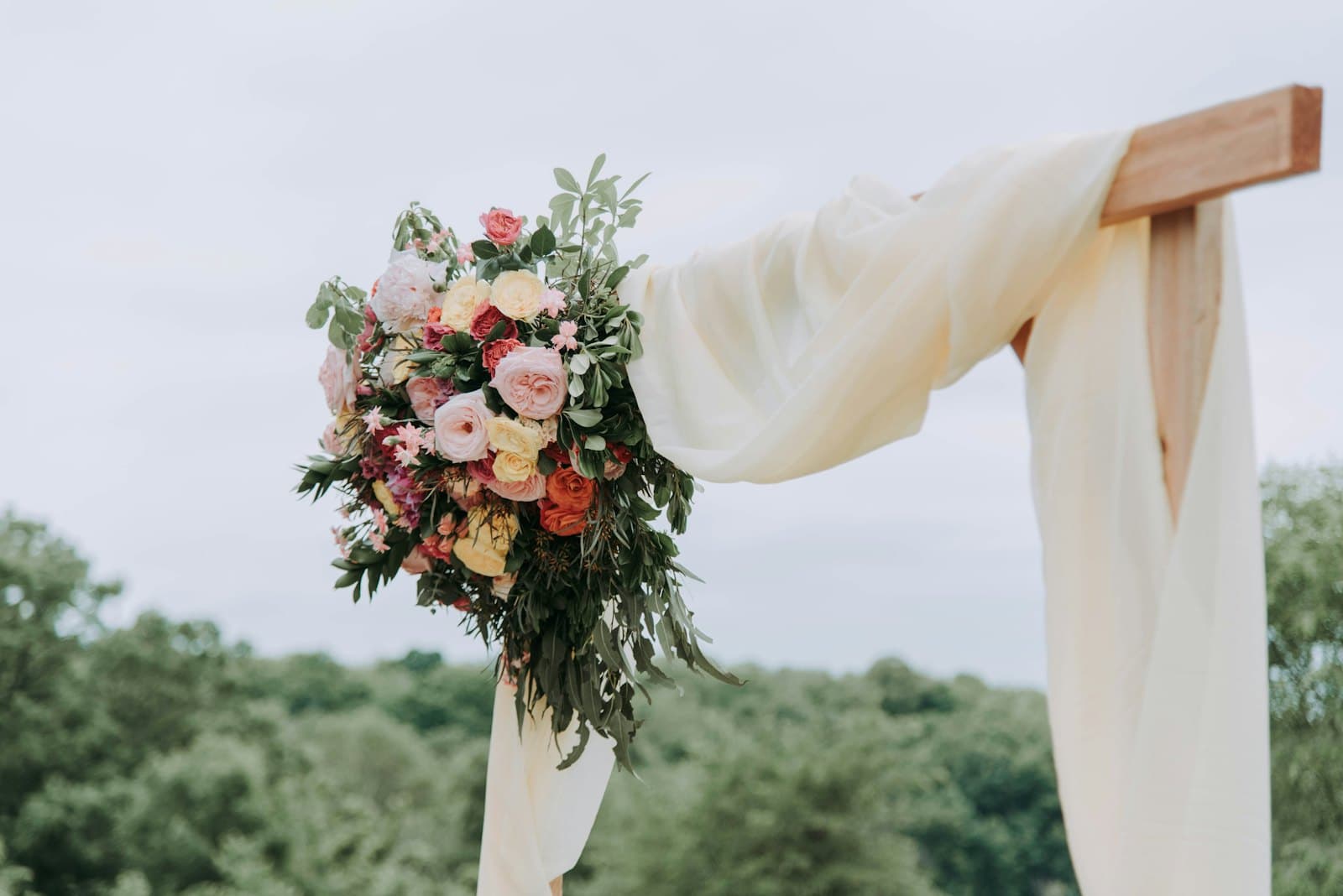 A wooden ceremony arch draped in ivory fabric and garden roses, green trees beyond