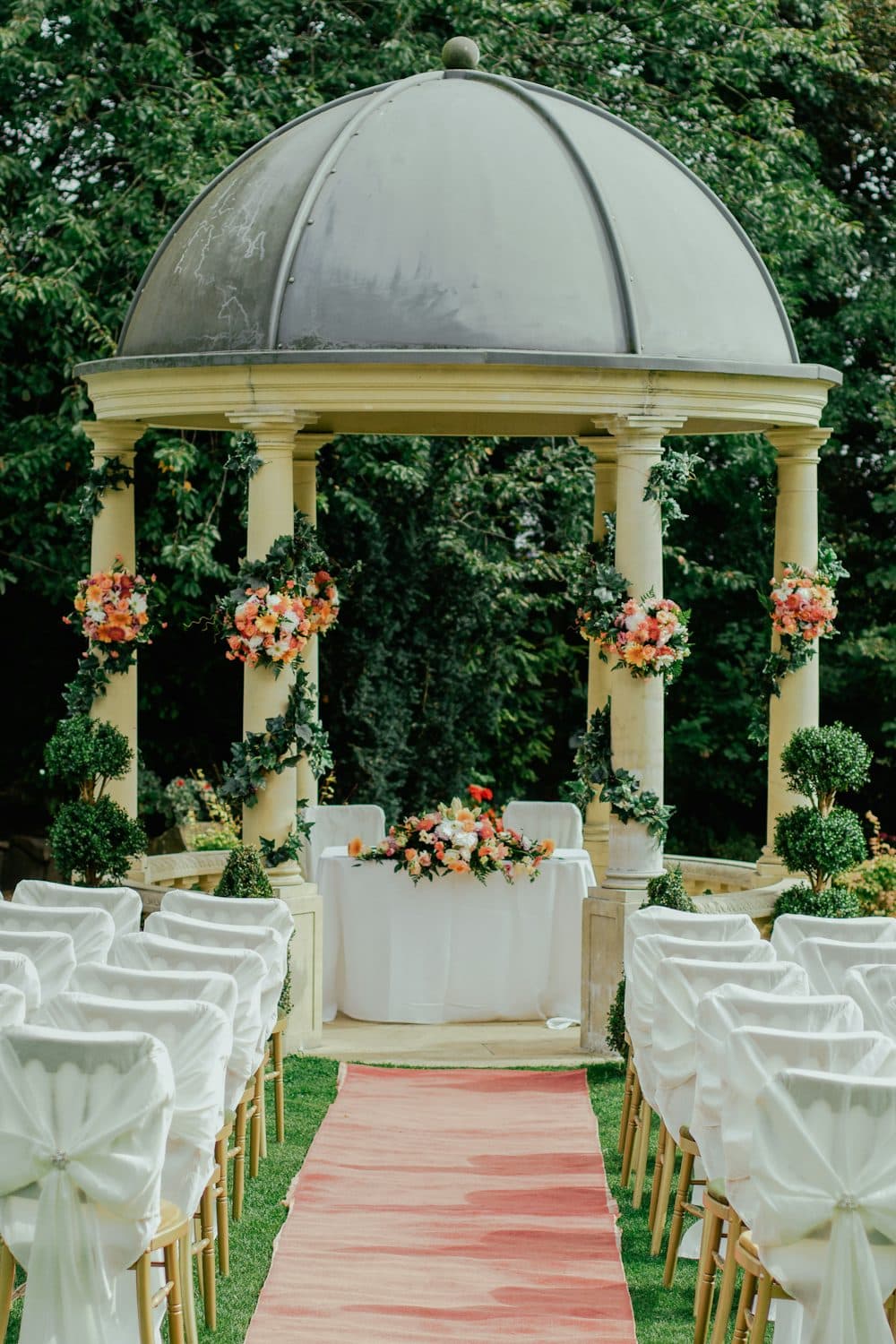 A garden pavilion dressed with flowers, rows of white-covered chairs along a rose-colored aisle
