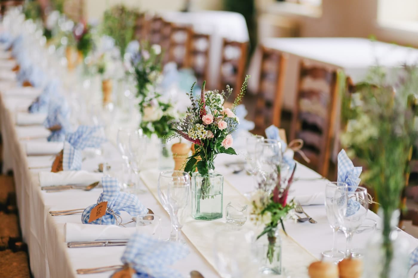 Long dinner tables set with white linen, gingham napkins and jars of wildflowers