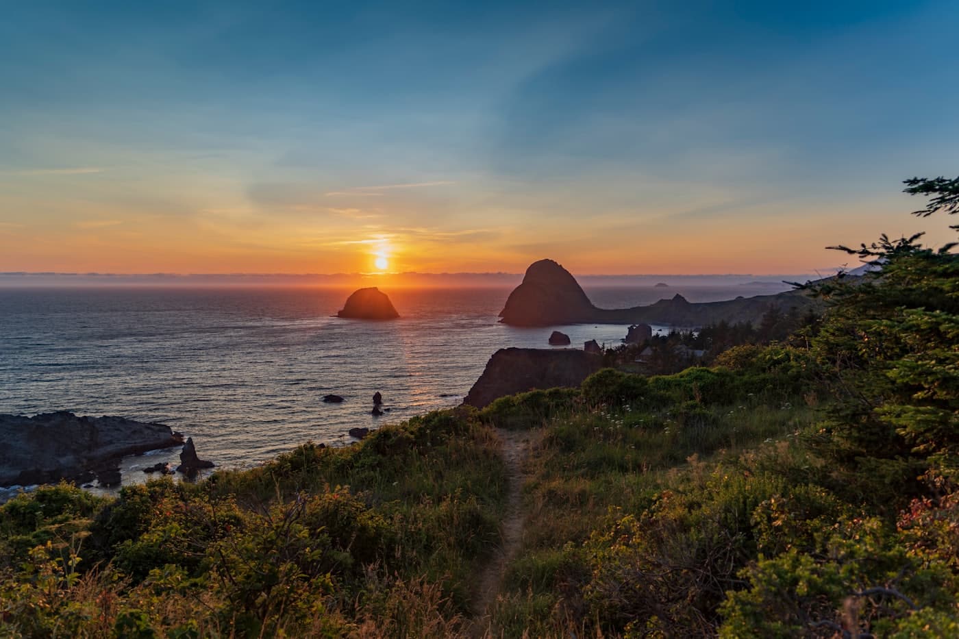 Sun setting behind offshore sea stacks, seen from a grassy headland trail