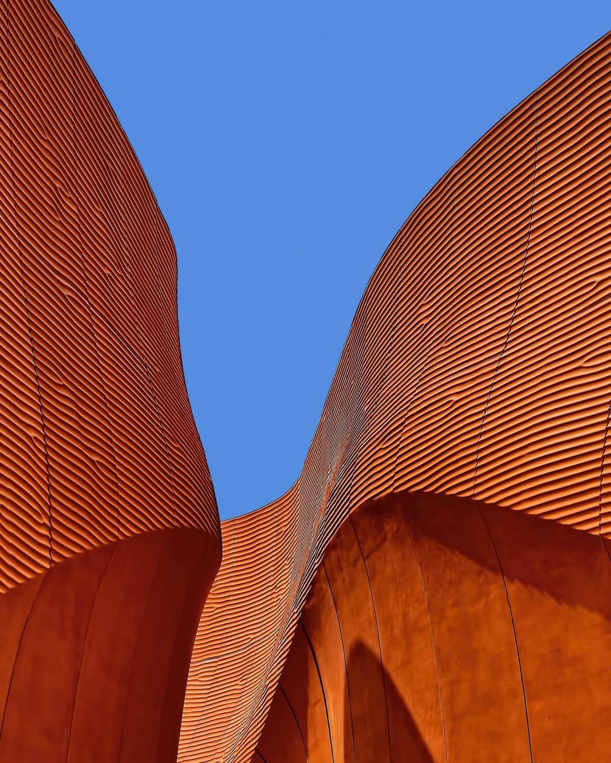 Curved corrugated corten-toned facade panels meeting against a clear blue sky