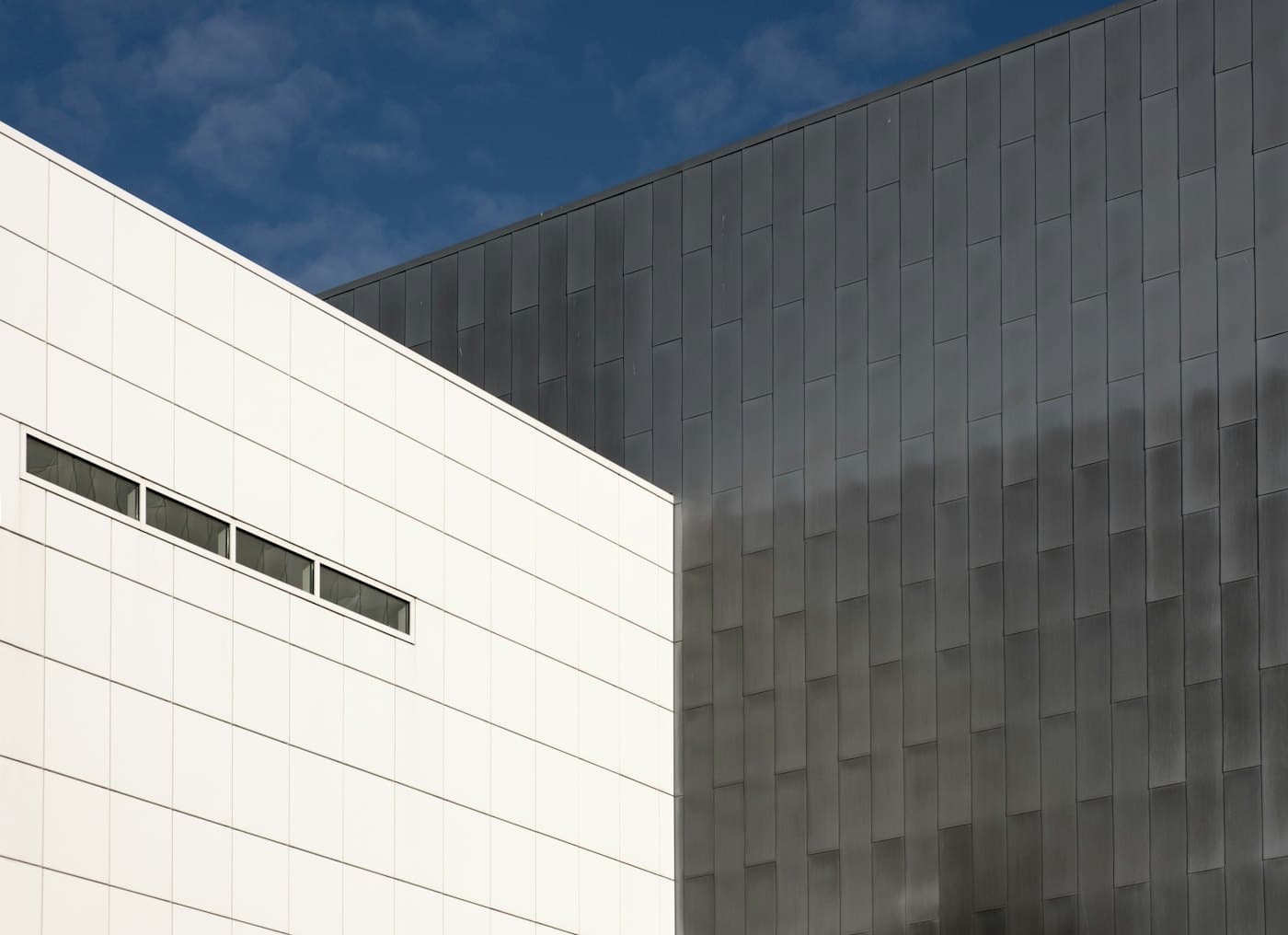 White tiled facade meeting a dark zinc-clad volume under a blue sky