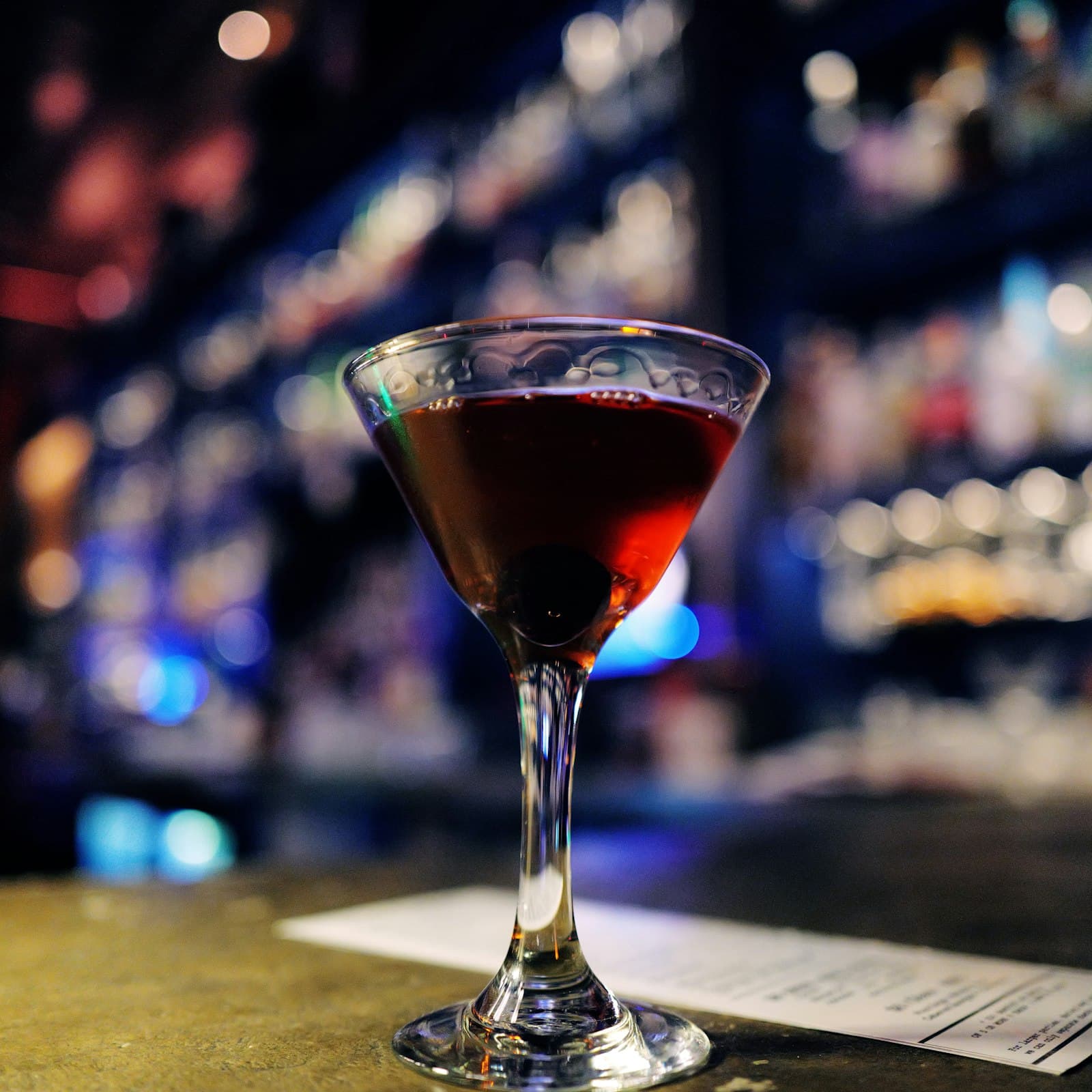 Dark red cocktail with a cherry in a stemmed glass on a bar, bottles glowing behind