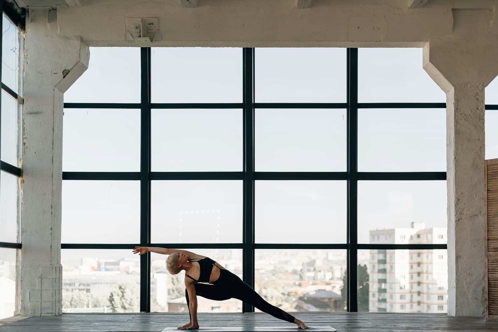 A student holding a long side-angle stretch in a bright loft studio with floor-to-ceiling windows