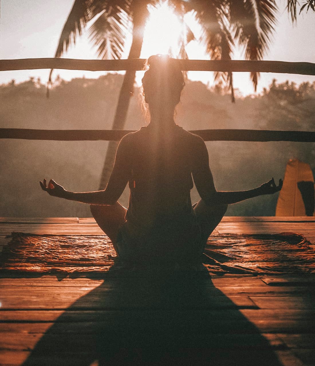 A seated meditation at sunrise, silhouetted against palm fronds on a wooden deck